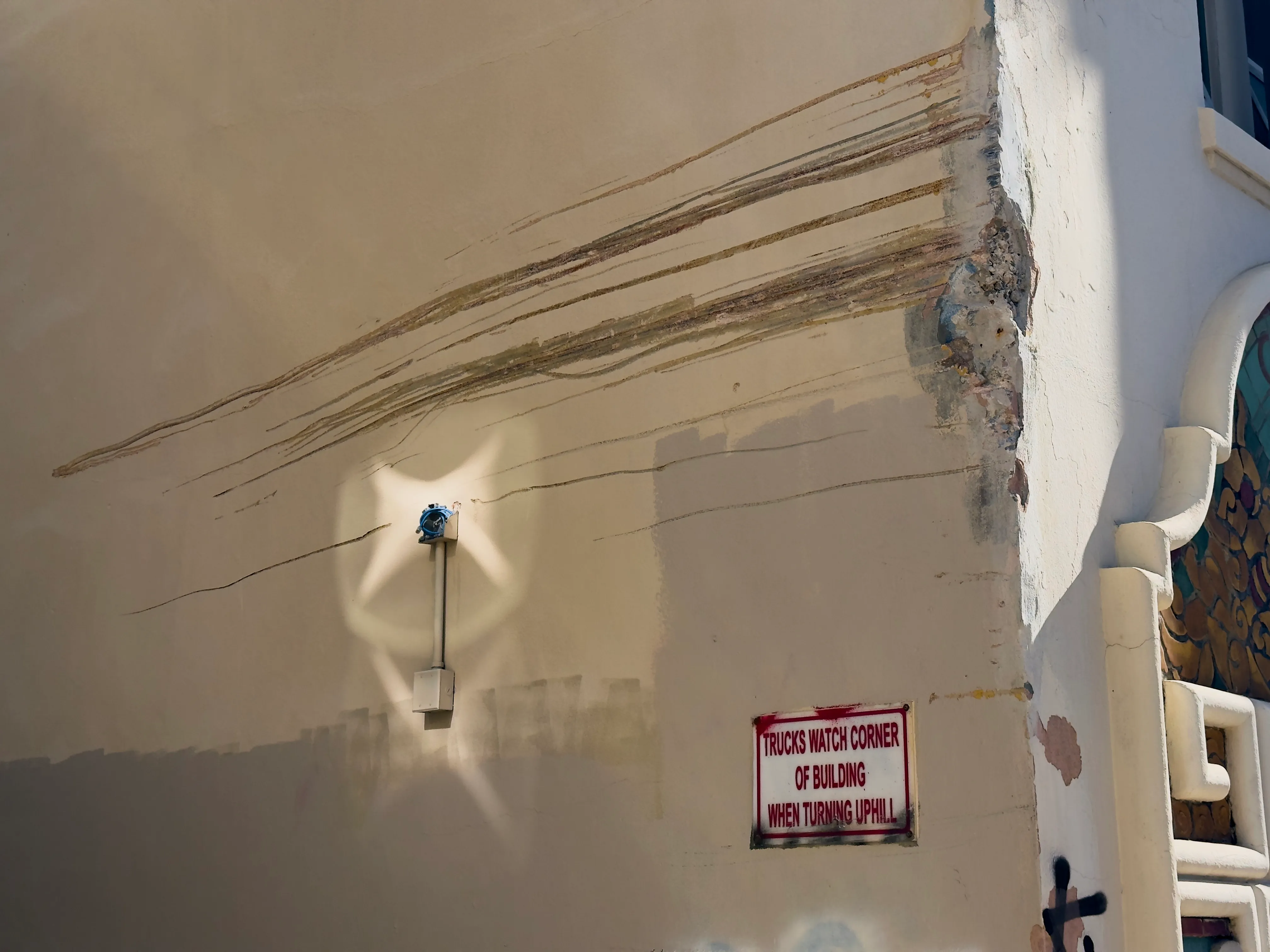 A sign that says "Trucks Watch Corner Of Building When Turning Uphill", right underneath a partially-destroyed side of a building...